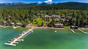 An aerial view of a lakeside resort with a marina, houses, and forested hills in the background. - Mh116 Mountain Harbor Cottage by the Lake (Whitefish)