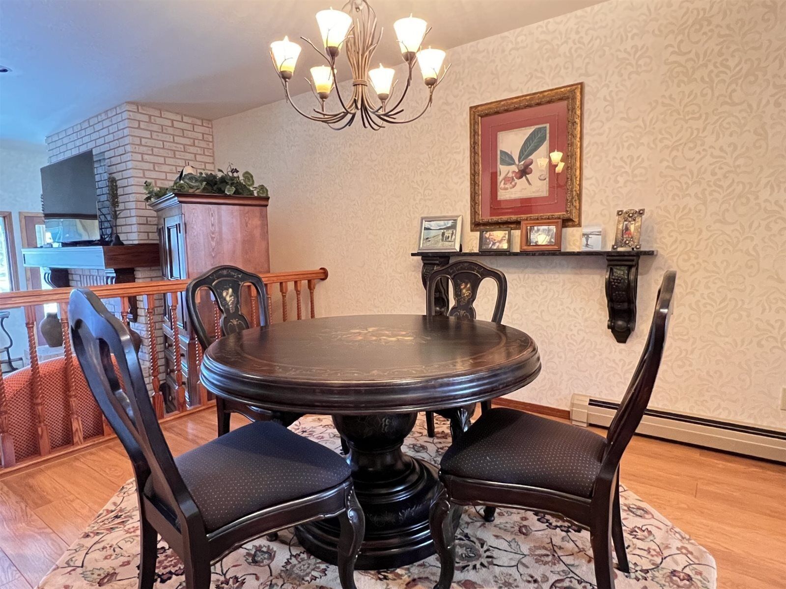 A dining area with a round table, four chairs, a chandelier, a fireplace, and a wall-mounted shelf with framed pictures.
