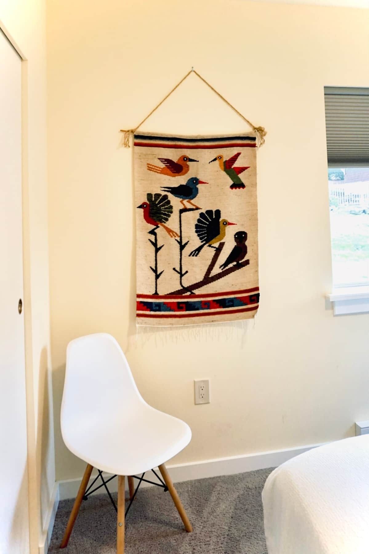 Een kamer met een witte stoel, wandtapijt met vogels en een raam met zonwering.