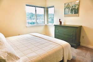 Une chambre avec un grand lit, une commode, une fenêtre donnant sur un port de plaisance et un tableau encadré accroché au mur.