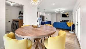 A modern kitchen with a central island, a dining area with a round wooden table and yellow chairs, and a living area with a blue sofa and yellow blanket. - Bellini House / Modern 3BD Near Dallas (Richardson)