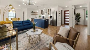 A spacious living area with a blue sofa, a coffee table, and a brown armchair. A kitchen with a central island and stainless steel appliances is visible in the background. - Bellini House / Modern 3BD Near Dallas (Richardson)