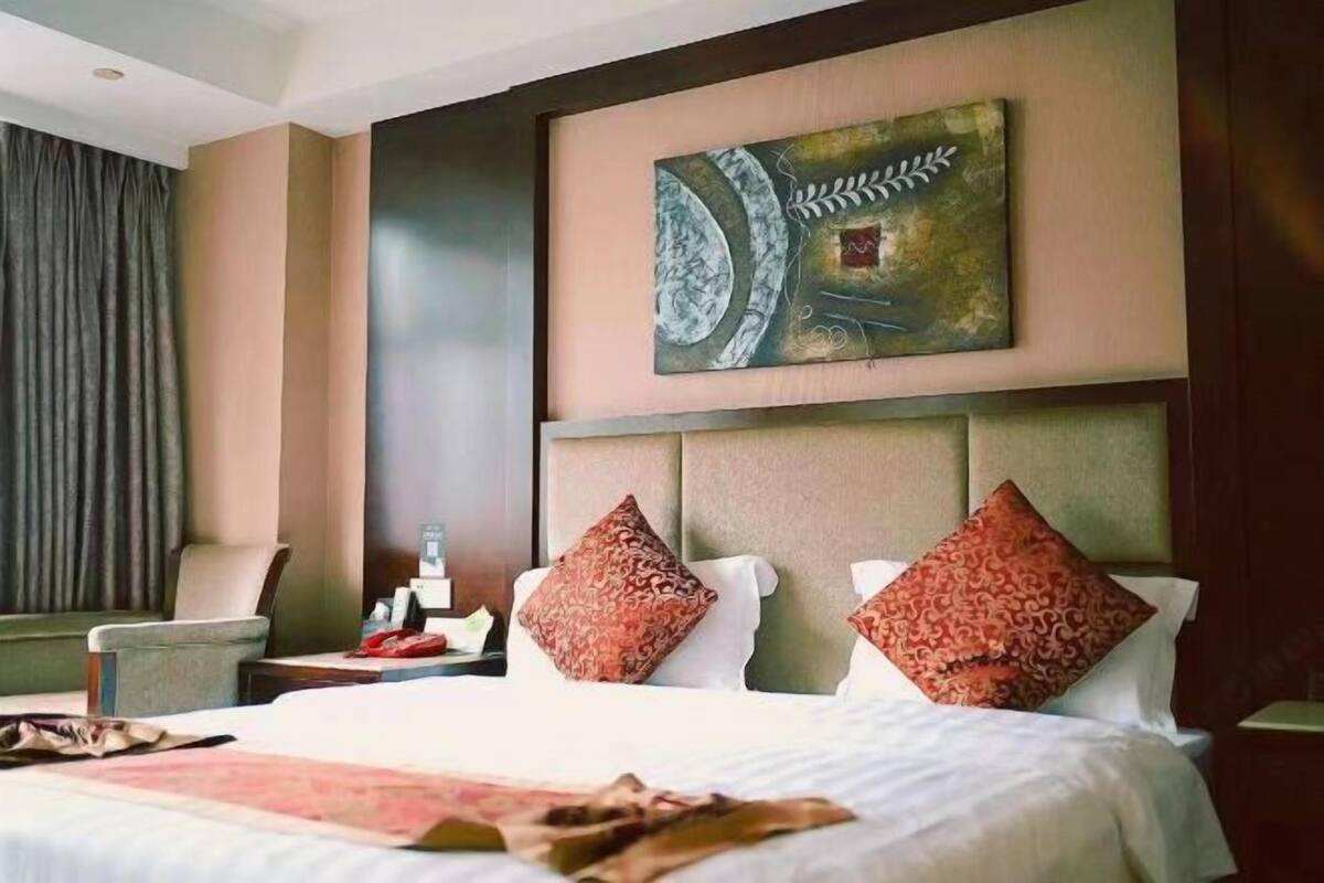 A hotel room with a large bed, two red patterned pillows, a headboard, a wall-mounted artwork, a nightstand, and a chair.