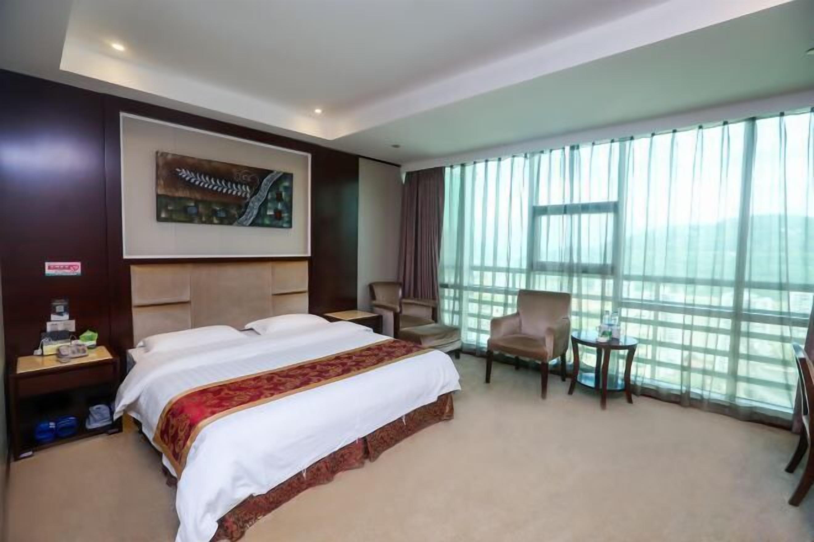 A hotel room with a large bed, a desk, a chair, and a view of the outdoors through large windows.