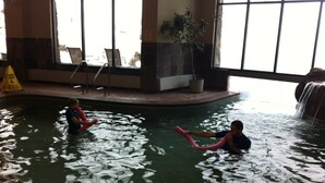 Uma piscina interna com duas pessoas na água, um sinal de segurança e uma planta ao fundo.
