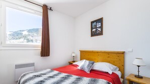 A hotel room with a bed, a window with a view of a town, a wooden headboard, a bedside table with a lamp, and a radiator.