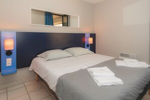 A hotel room with a bed, two bedside lamps, a window with curtains, and a small table with towels.