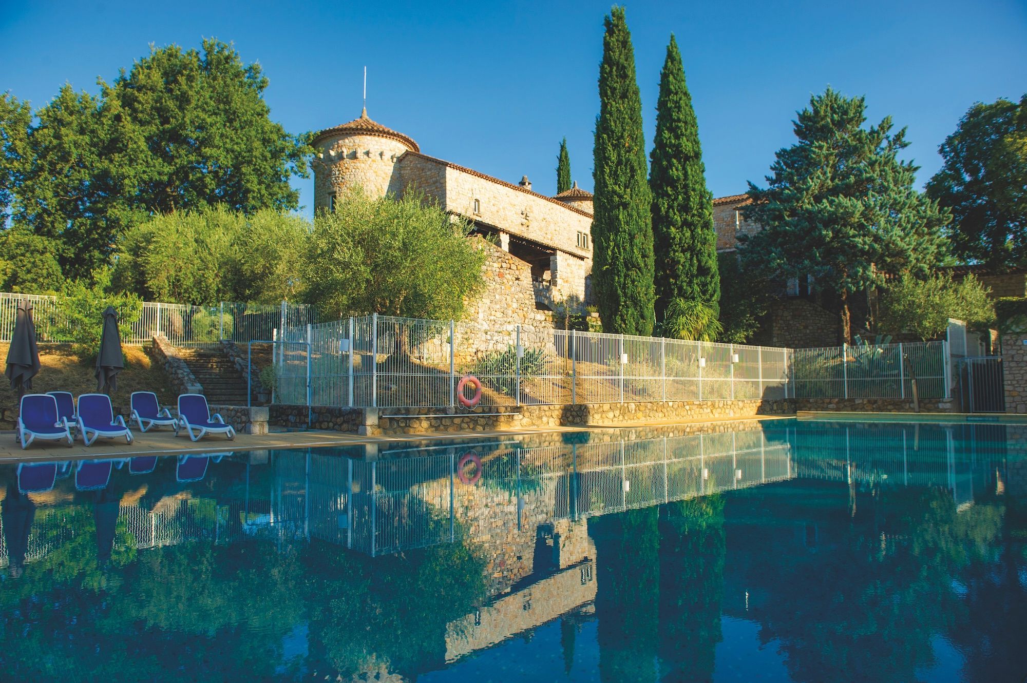 A swimming pool with lounge chairs, a stone building with a tower, and tall cypress trees.