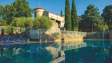 A swimming pool with lounge chairs, a stone building with a tower, and tall cypress trees.