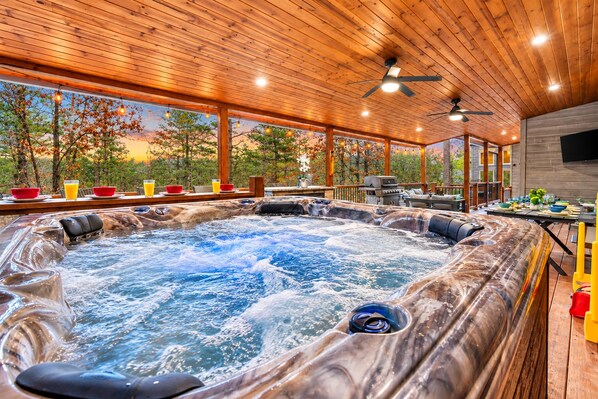 A hot tub with a view of trees and a sunset, wooden ceiling, and a dining area with outdoor seating.