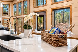 A kitchen with a marble countertop, a sink, a potted orchid, a wicker basket with kitchen utensils, and a view of a forest through large windows.