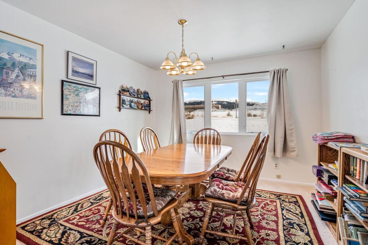 A dining area with a wooden table and chairs, a chandelier, a bookshelf, and a window with a view of the outdoors.