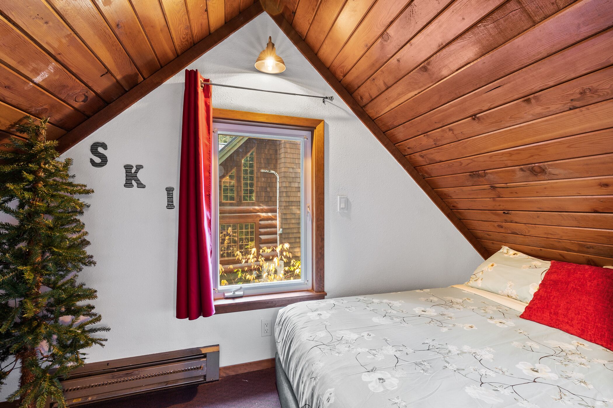 A bedroom with a sloped ceiling, a window with red curtains, a bed with floral bedding, and a small tree in the corner.