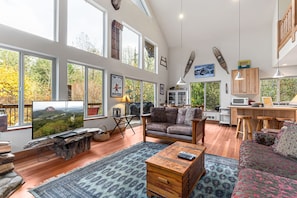 A spacious living area with a large TV, wooden furniture, and a view of trees through large windows.