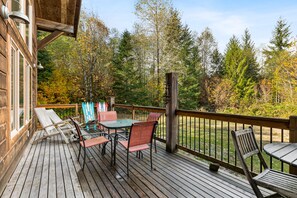 A wooden deck with outdoor furniture, including a table and chairs, overlooking a forested area with autumn foliage.
