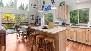 Une cuisine moderne avec des meubles en bois, un grand îlot central doté de tabourets, des luminaires suspendus et une vue sur les arbres à travers les fenêtres.