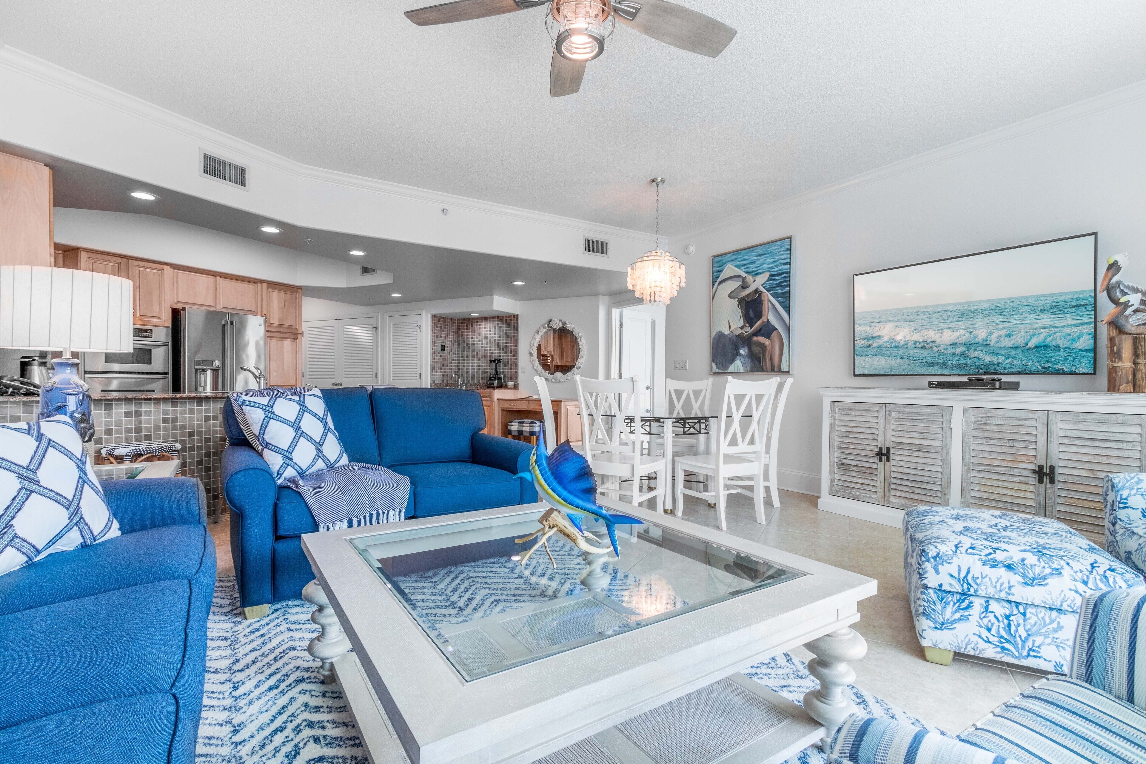 Uma sala de estar moderna com um sofá azul, uma mesa de centro de vidro e uma área de jantar com cadeiras brancas.