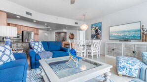 A modern living room with a blue sofa set, a glass coffee table, and a dining area with white chairs. - Nautical Two Bedroom Condo Overlooking the Perdido Bay and Pools! Complimentary Boat Slip! (Orange Beach)