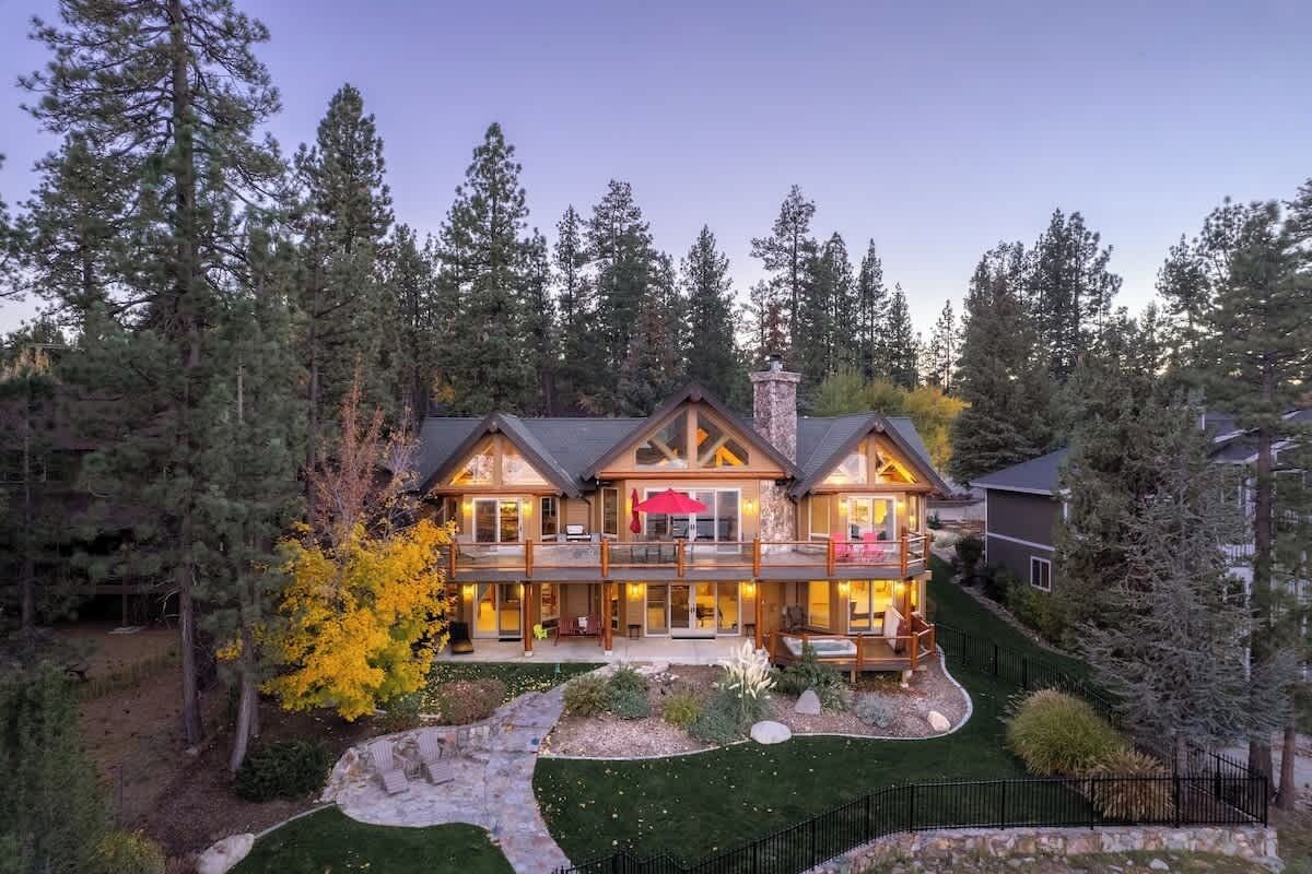 A multi-story log cabin with a deck, surrounded by a forest.