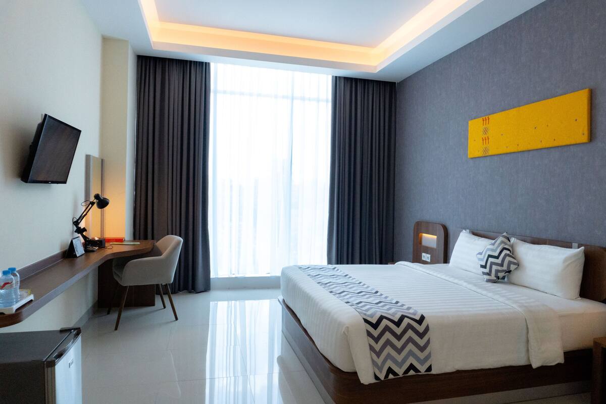 A hotel room with a bed, a desk with a chair, a television, and a large window with curtains.