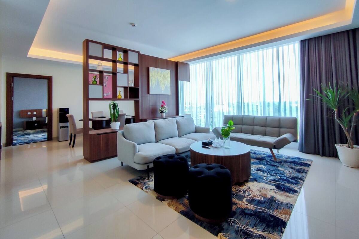 A modern living room with a sofa, coffee table, and ottoman, a desk with a computer, and a large window with curtains.