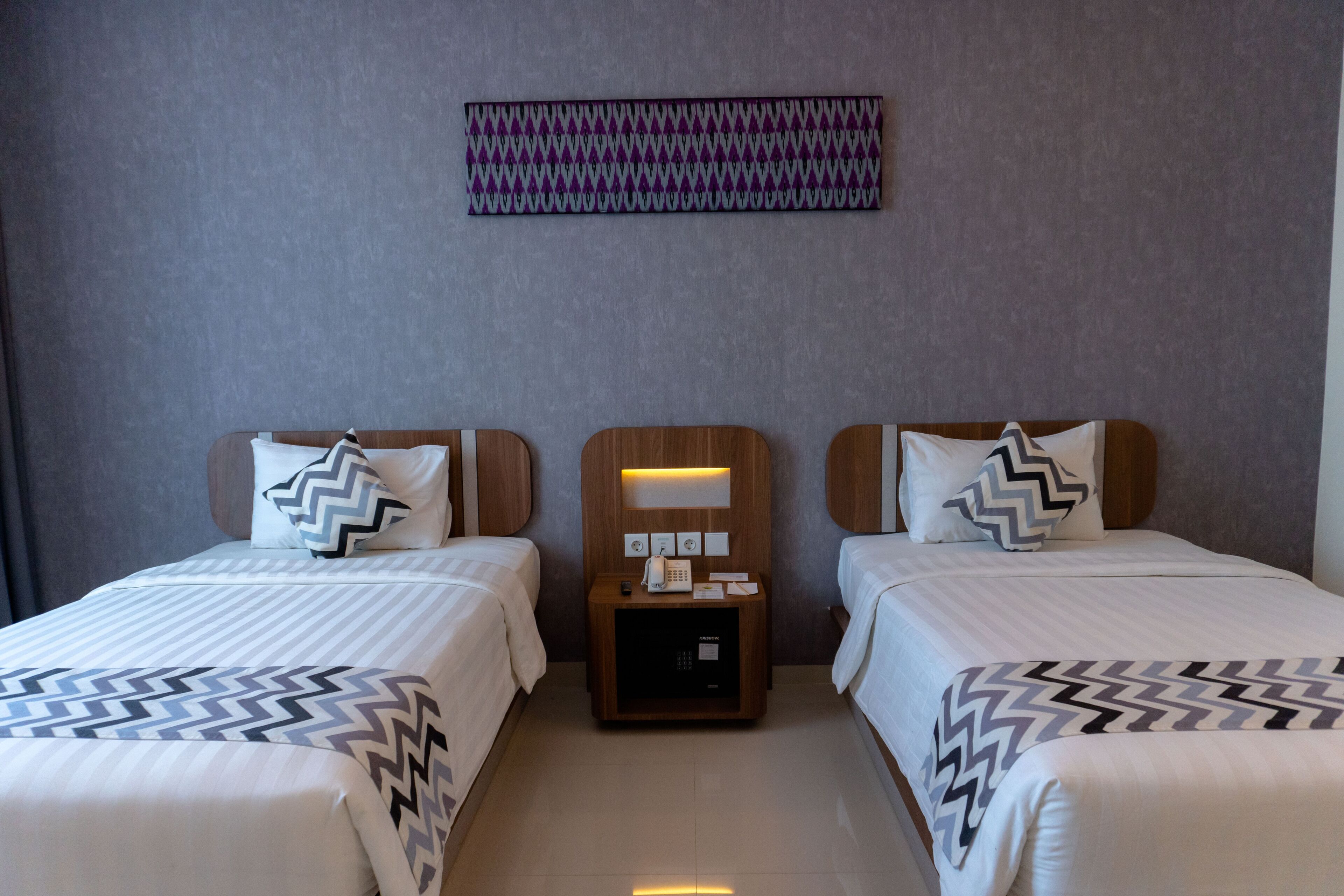 A hotel room with two beds, each with a patterned bedspread, a nightstand with a phone and a small table, and a wall-mounted light fixture.