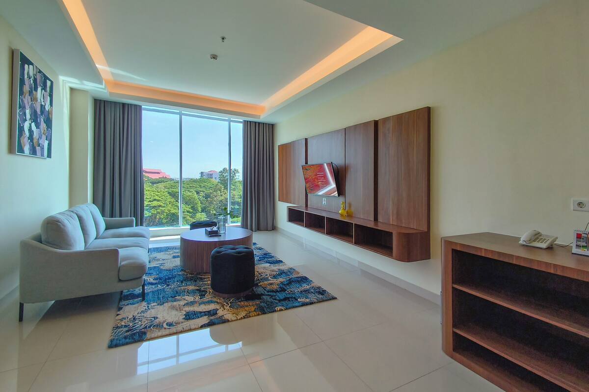 A modern living room with a sofa, a coffee table, a TV mounted on the wall, and a large window with curtains.