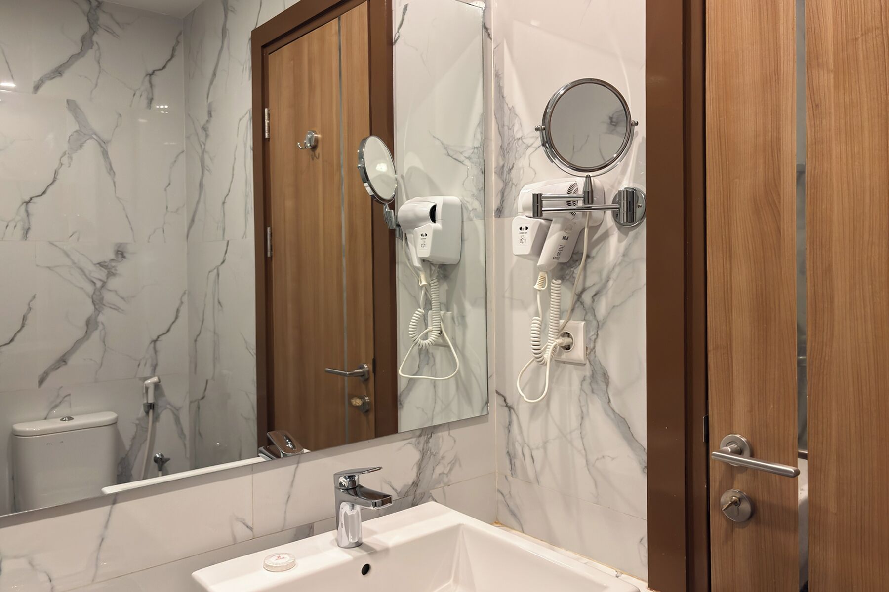 A bathroom with a marble wall, a sink, a mirror, and a hairdryer mounted on the wall.