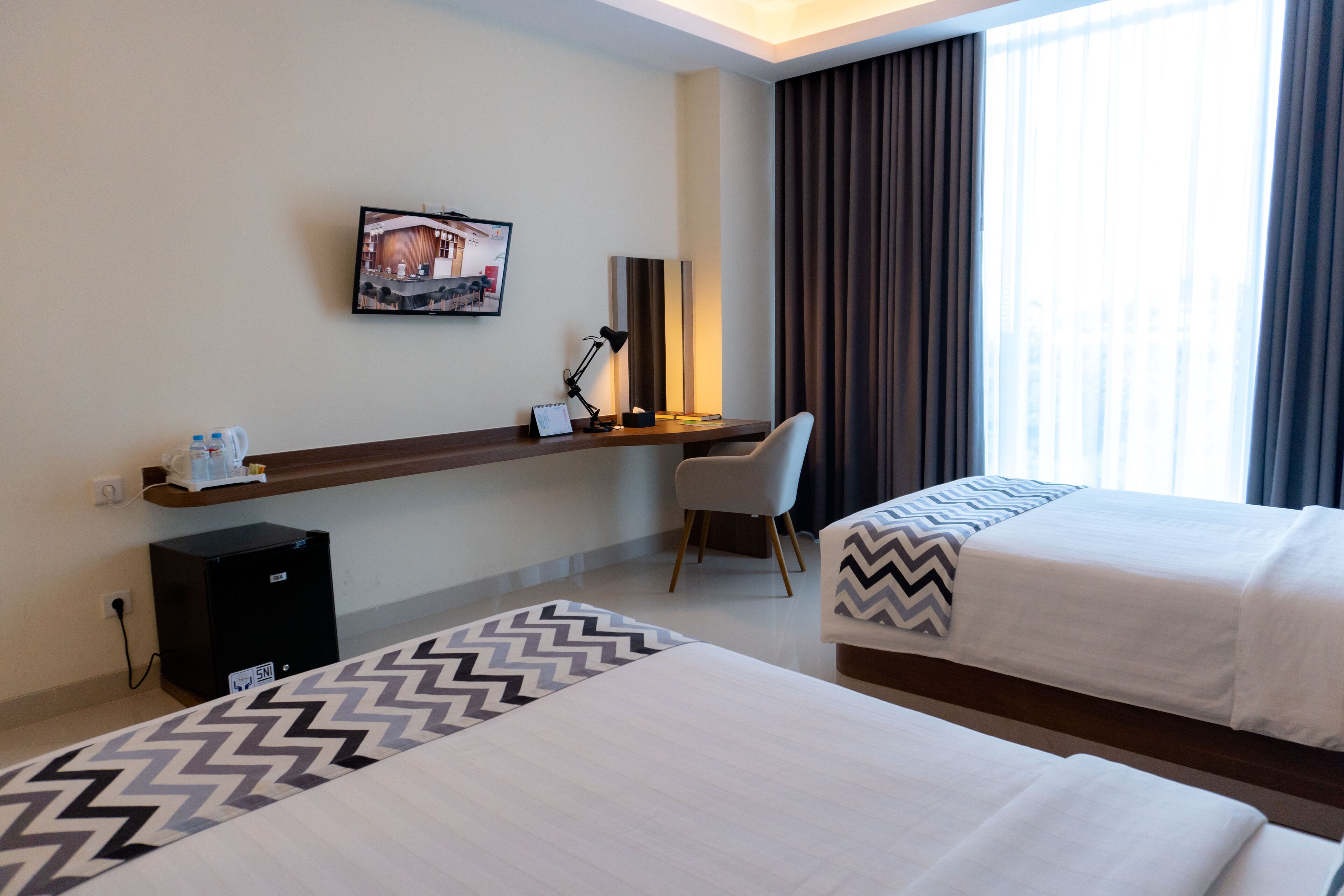A hotel room with two beds, a desk with a chair, a small refrigerator, a lamp, and a picture on the wall.