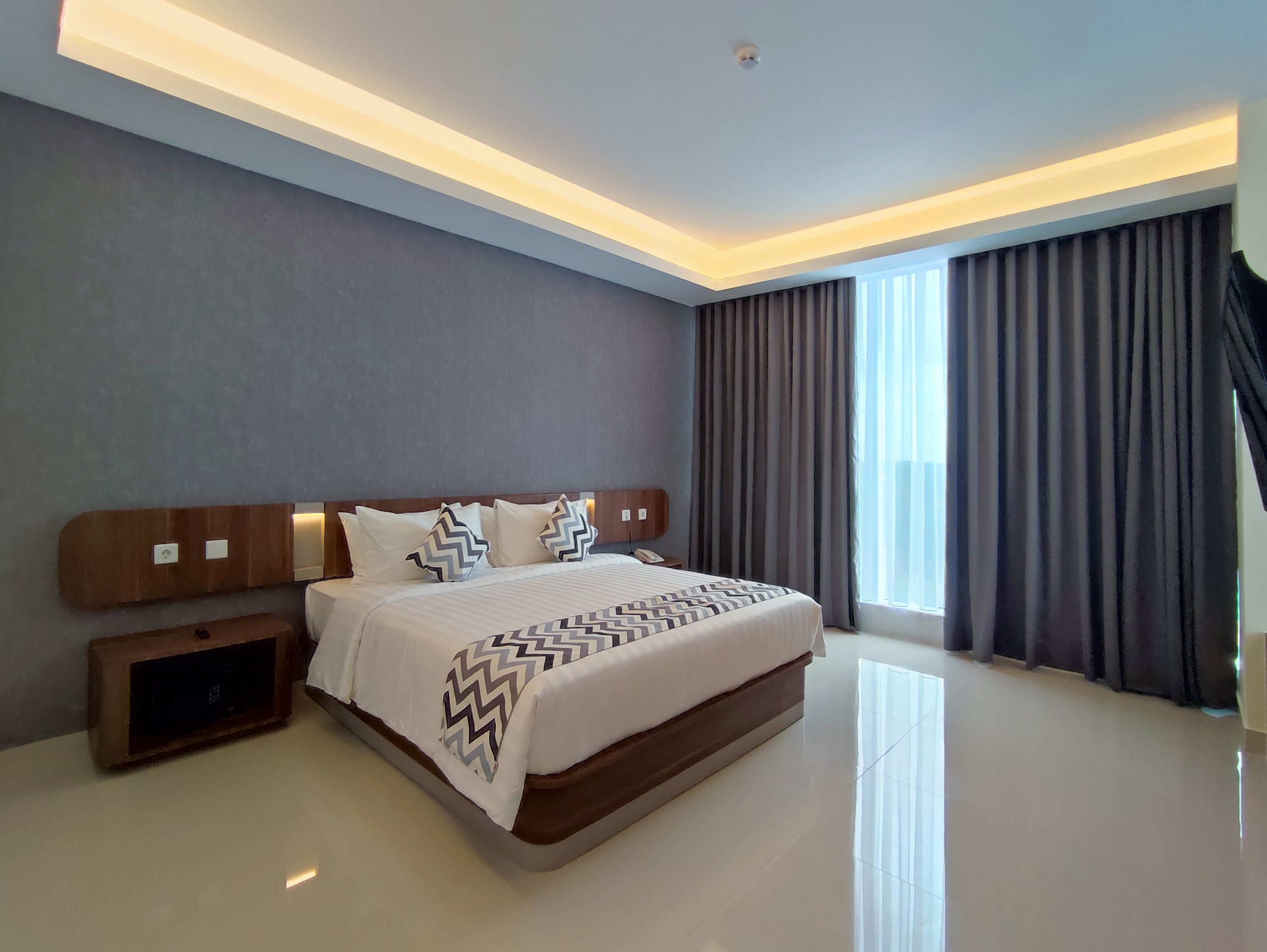 A modern hotel room with a large bed, a wooden headboard, a nightstand, and a large window with curtains.