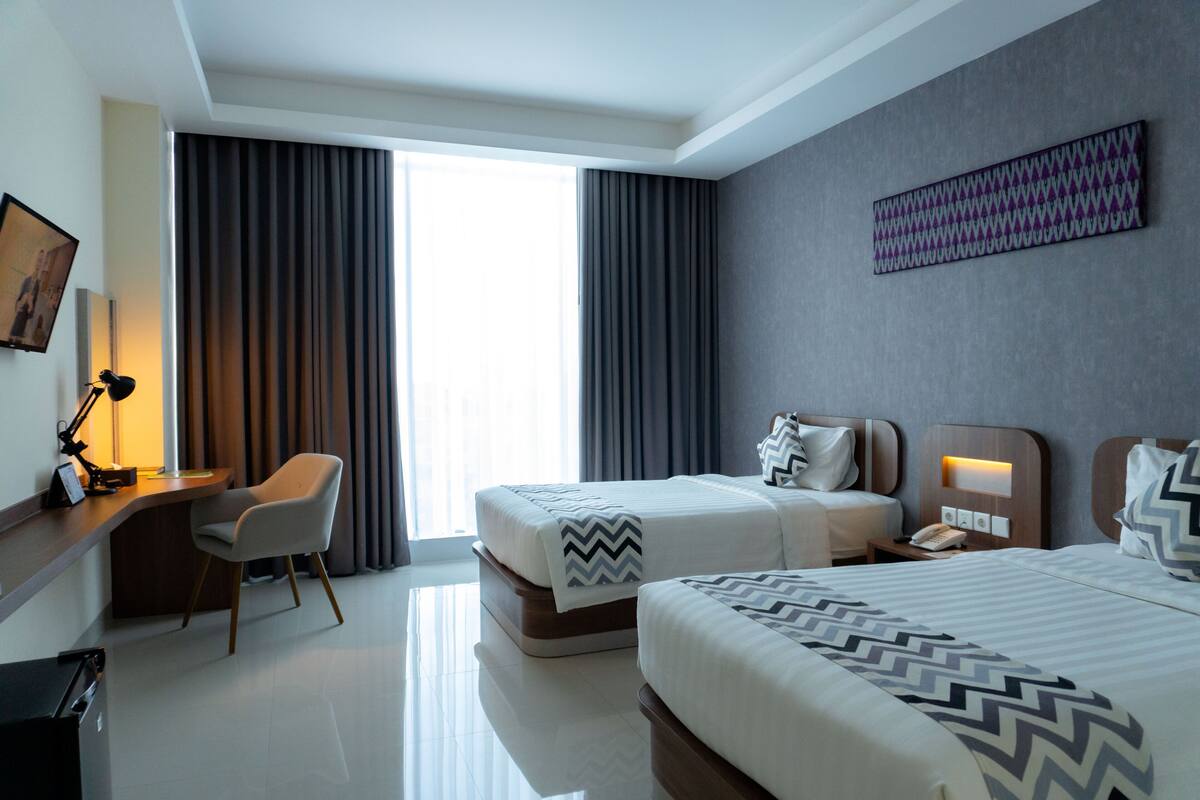 A hotel room with two beds, a desk, a chair, and a large window with curtains.