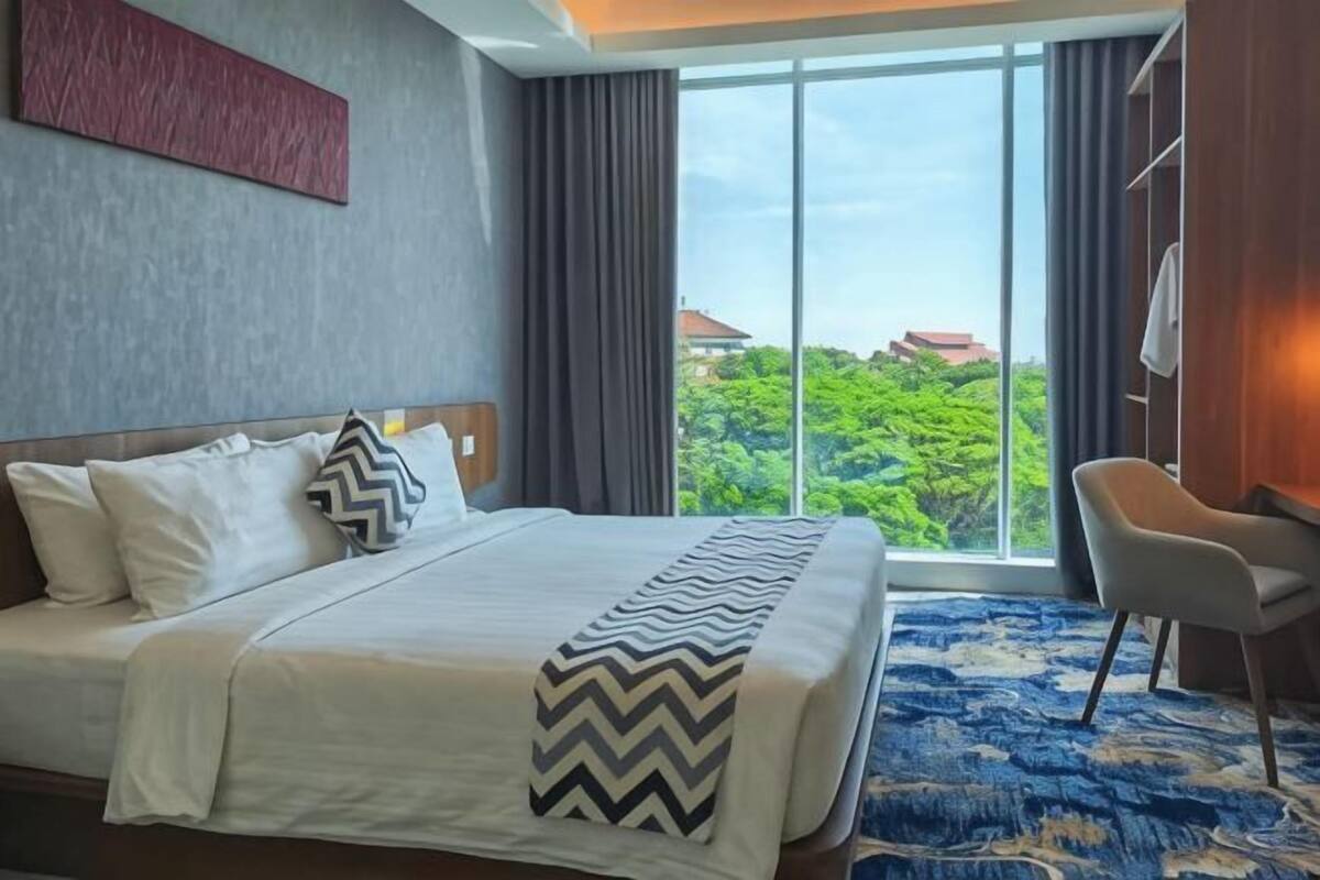 A hotel room with a large bed, a chair, a view of greenery, and a window.
