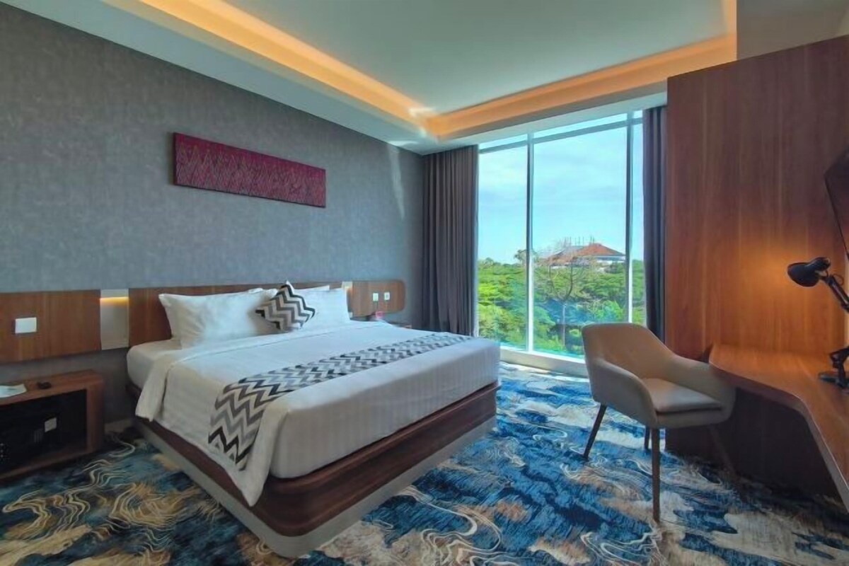Une chambre d’hôtel avec un grand lit, un bureau et une chaise. Il y a une fenêtre donnant sur un espace verdoyant et un immeuble.