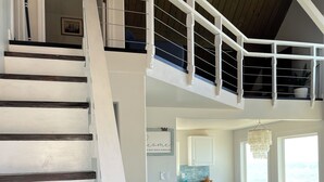 A spacious living area with a high ceiling, a staircase with dark wooden steps, and a view of the kitchen and dining area. - West Beach Bungalow: Whidbey Waterfront Beach House VIEWS + 5 Star Reviews (Oak Harbor)