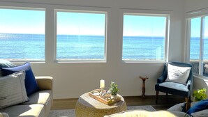 A room with large windows overlooking the sea, a sofa, a chair, and a small table with decorative items. - West Beach Bungalow: Whidbey Waterfront Beach House VIEWS + 5 Star Reviews (Oak Harbor)