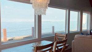 A dining area with a wooden table and chairs, large windows with a view of the sea, and a hanging light fixture. - West Beach Bungalow: Whidbey Waterfront Beach House VIEWS + 5 Star Reviews (Oak Harbor)