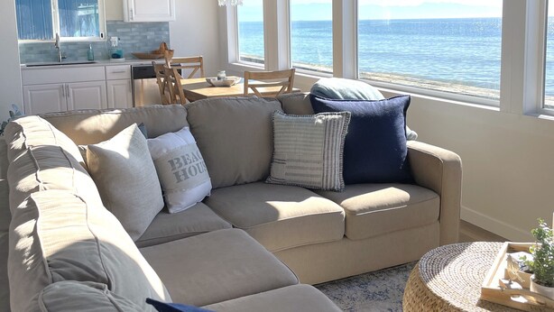 A spacious living area with a large beige sectional sofa, a round woven coffee table, and a view of the sea through large windows. - West Beach Bungalow: Whidbey Waterfront Beach House VIEWS + 5 Star Reviews (Oak Harbor)