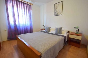 A bedroom with a bed, two bedside tables, a nightstand, a lamp, a window with curtains, and a wall-mounted picture.