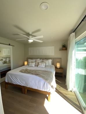 Quarto com uma cama grande, um ventilador de teto, abajures, janela com cortinas e vista para o verde lá fora.