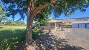 A residential property with a covered patio, a gravel driveway, and a large tree providing shade. - Quaint Farmhouse on  Palisade Peach Orchard close to Wineries (Palisade)