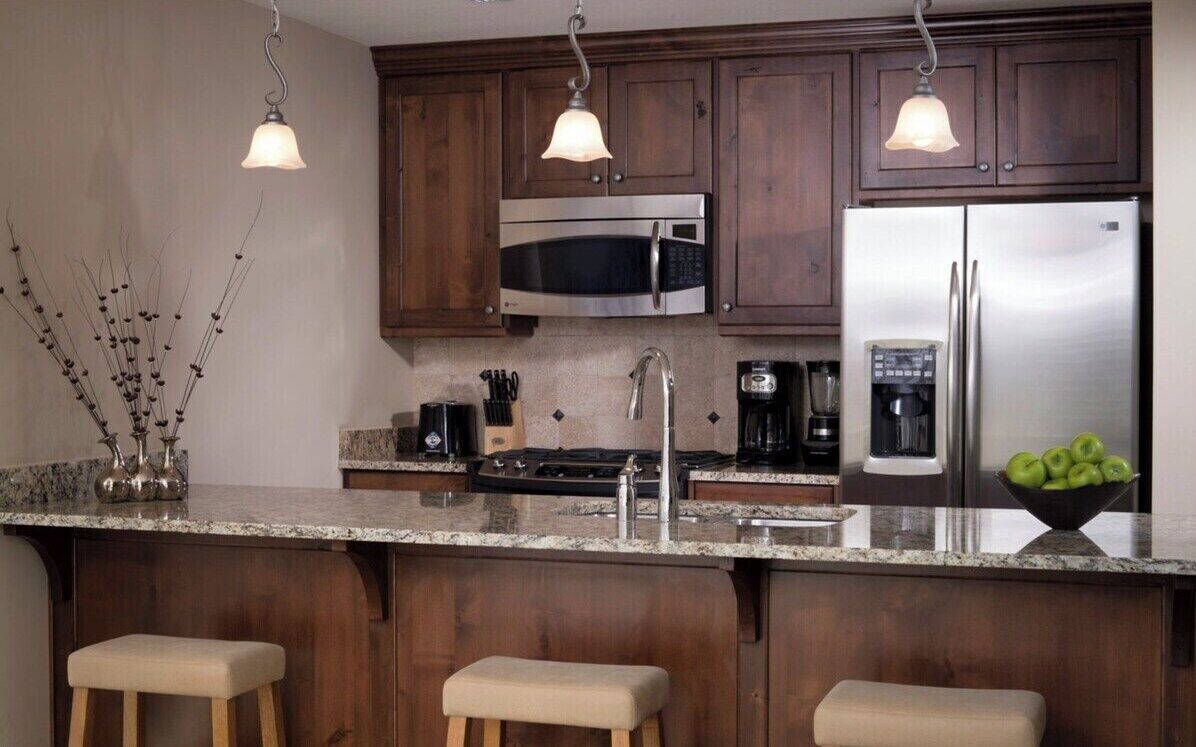 A kitchen with dark wooden cabinets, a stainless steel refrigerator, a microwave, a coffee maker, and a bowl of green apples on the counter.