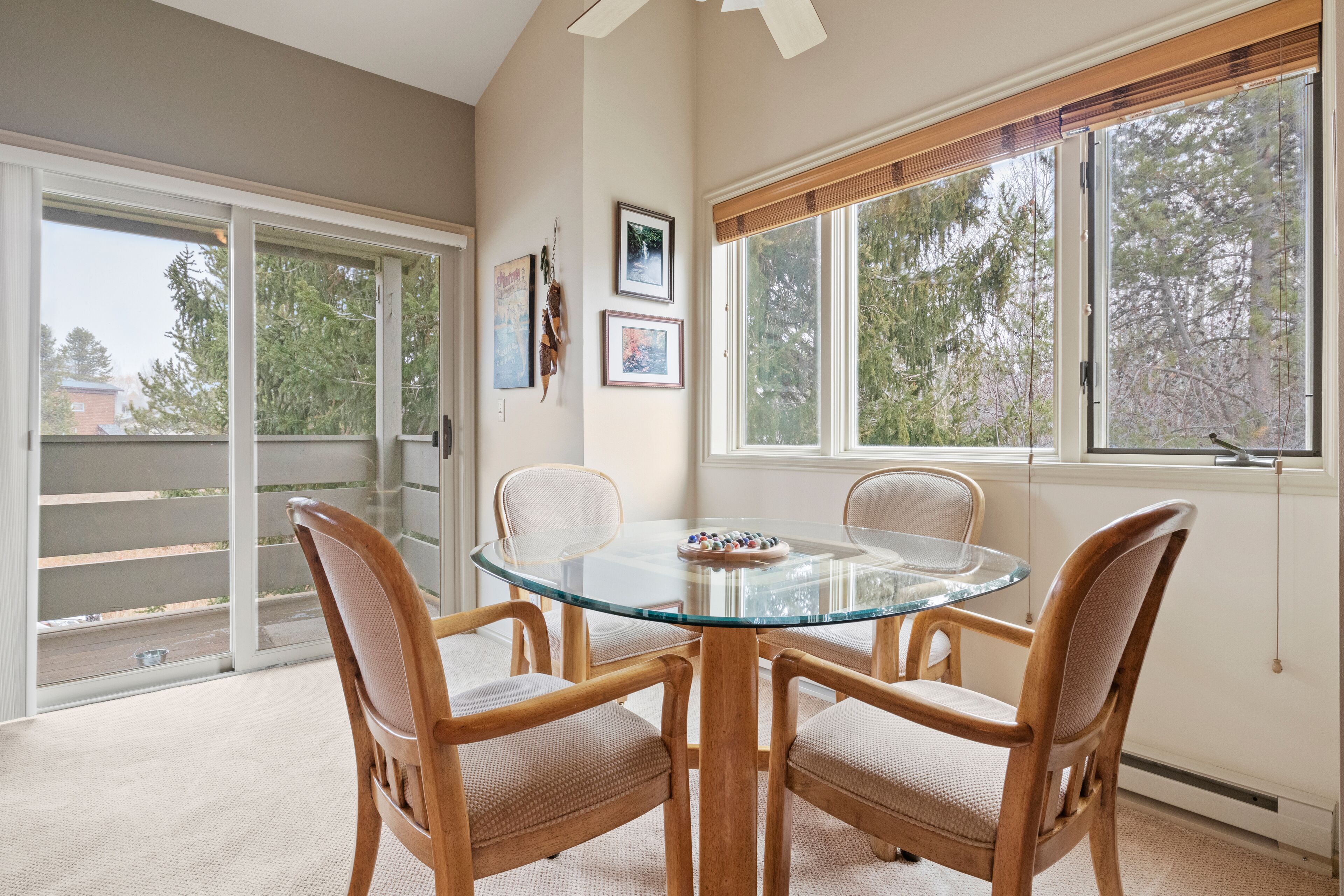 Un coin repas avec une table en verre entourée de chaises en osier, de grandes fenêtres équipées de stores et une vue sur les arbres à l’extérieur.