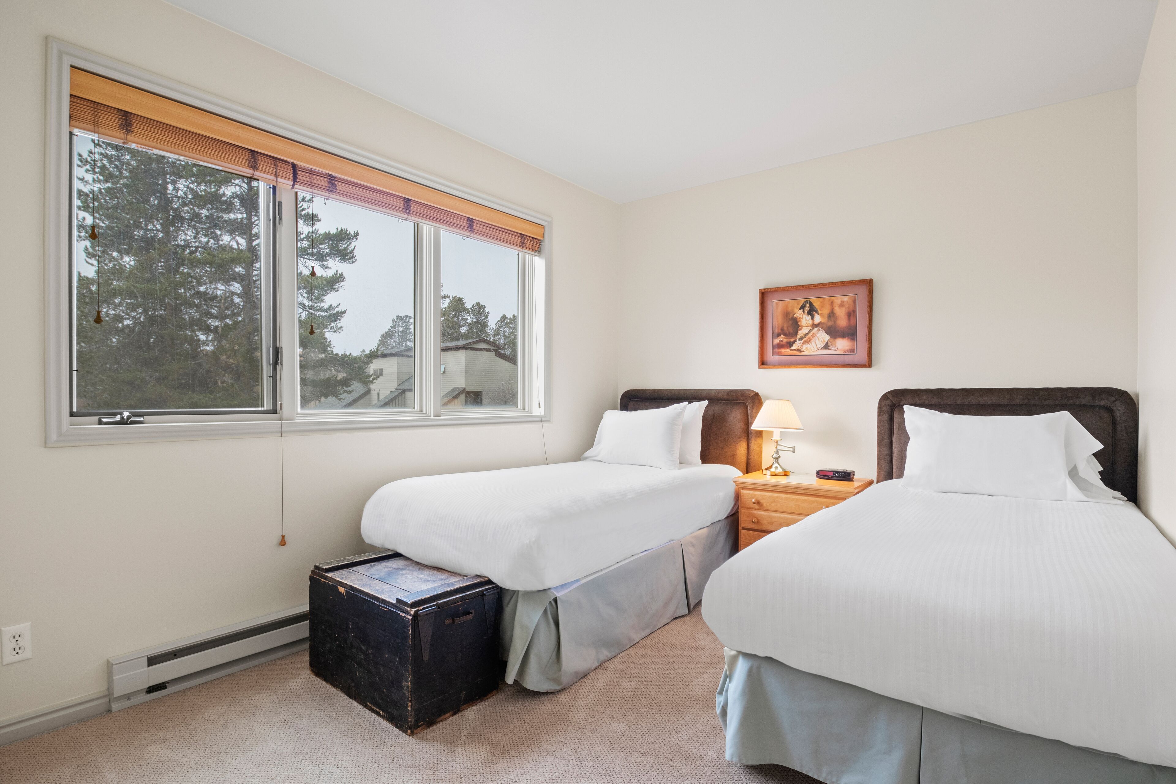 A room with two beds, a window with blinds, a framed picture on the wall, and a small wooden nightstand with a lamp.