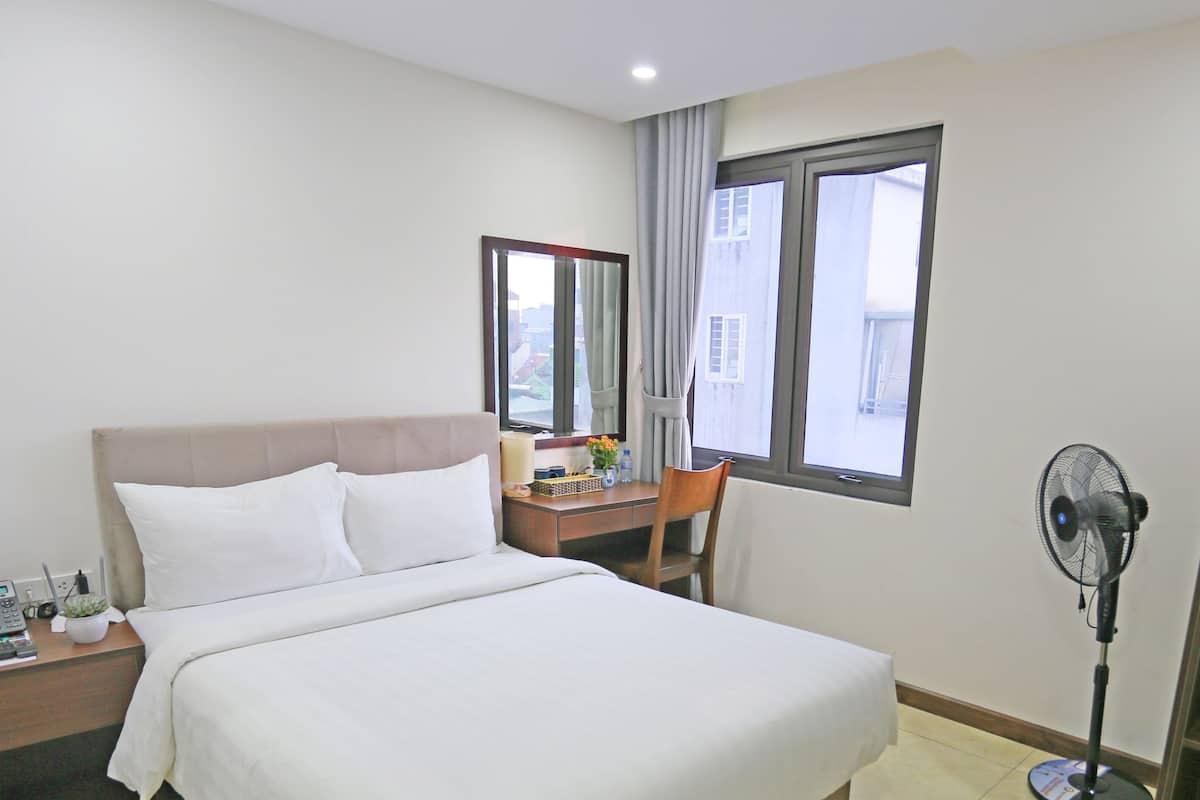 Una habitación de hotel con cama, mesita de noche, escritorio, silla y un ventilador de pie.
