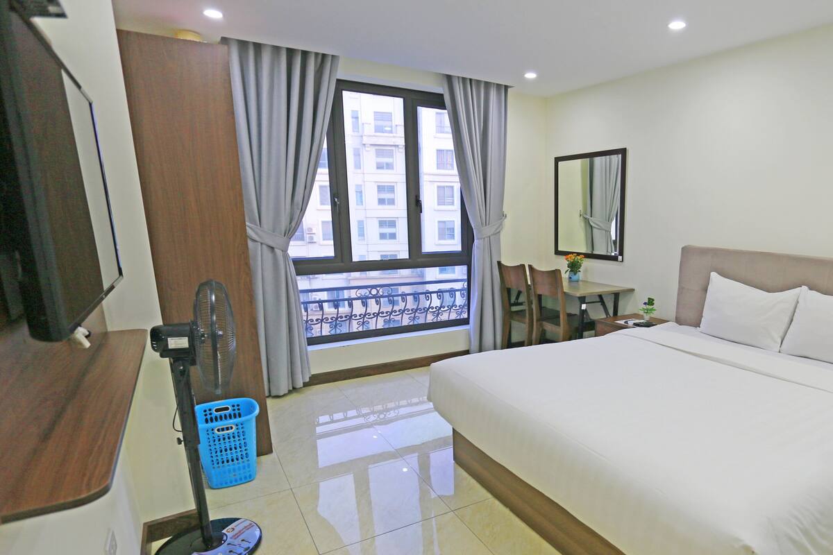 Habitación de hotel con una cama grande, un escritorio con una silla, una mesita con un jarrón de flores, una ventana con vista a edificios y un ventilador en el suelo.