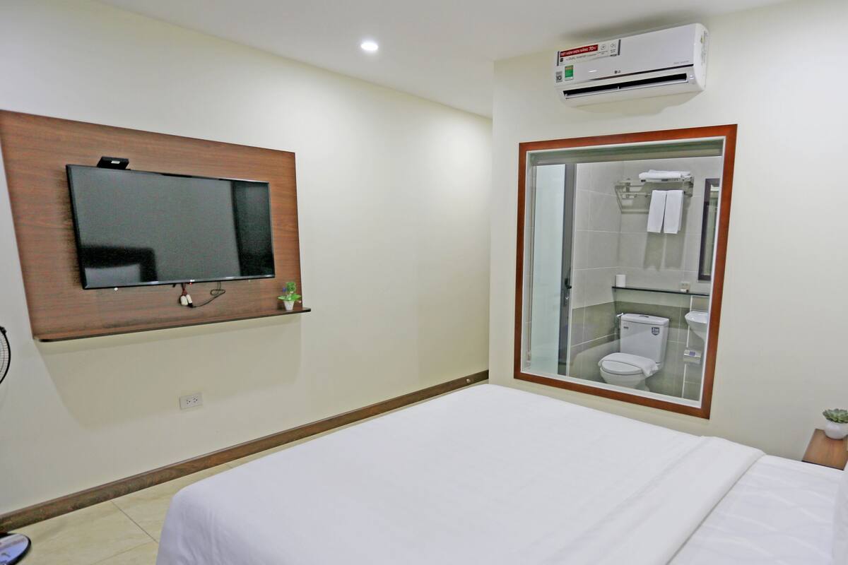 Habitación de hotel con una cama, un televisor montado en la pared, un equipo de aire acondicionado y un baño visible a través de una puerta de cristal.