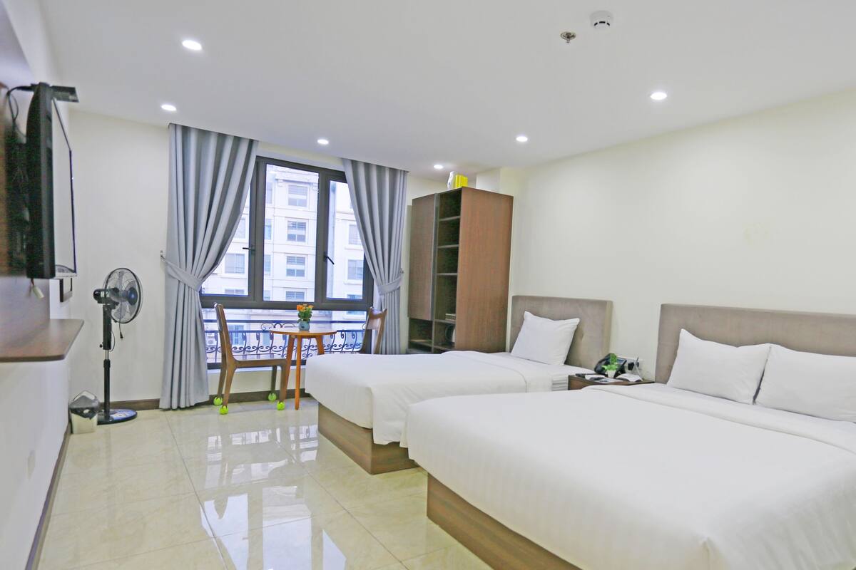 Habitación de hotel con dos camas, un escritorio, una silla, una ventana con cortinas, un ventilador y vistas a edificios.