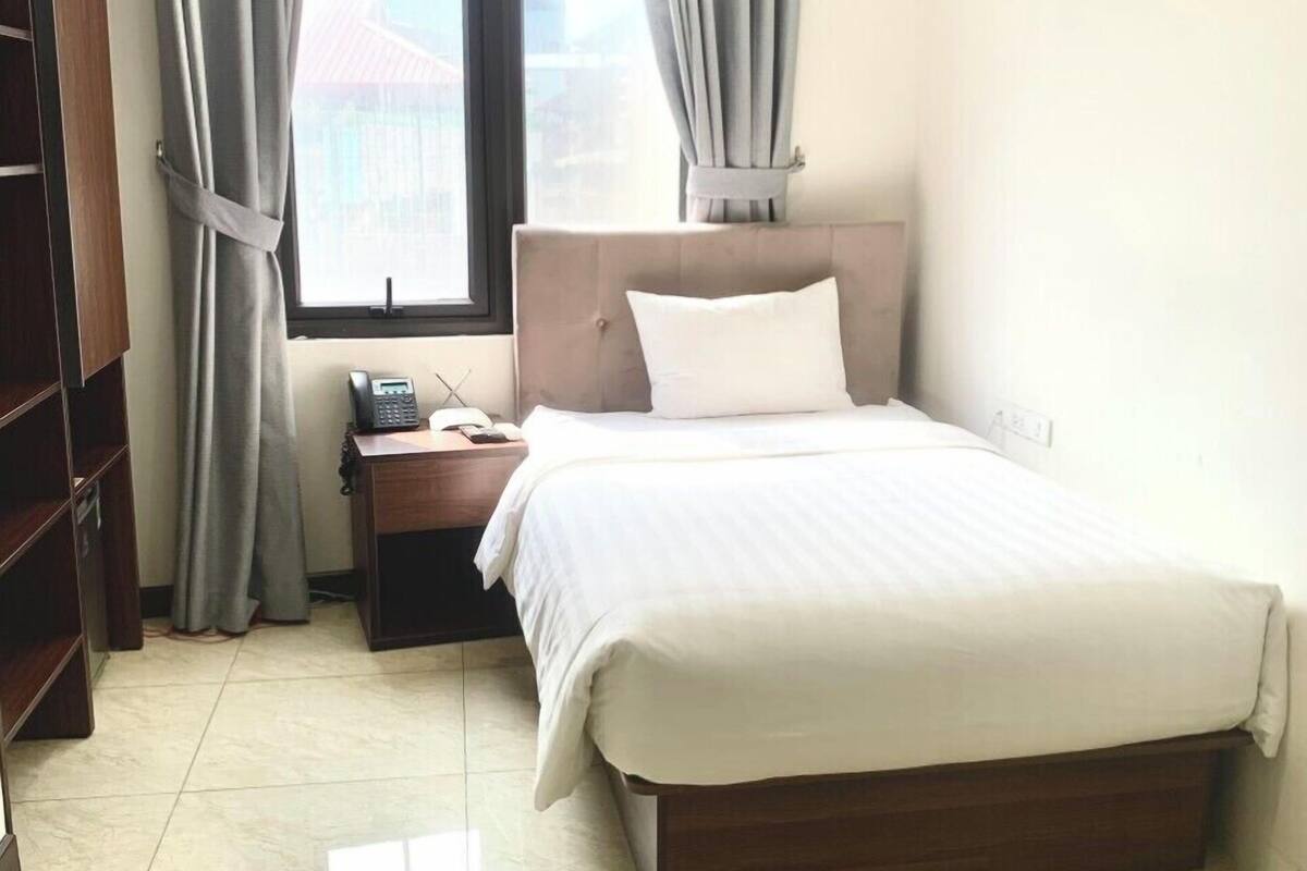 Una habitación de hotel con cama, mesita de noche, teléfono y ventana con cortinas.