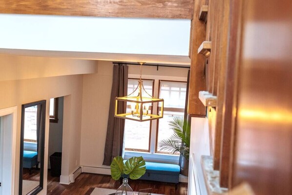 A high-ceilinged room with a wooden floor, a hanging geometric light fixture, and a view of a blue bench outside a window.