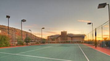 An outdoor tennis court with a net, surrounded by a fence, and street lamps.